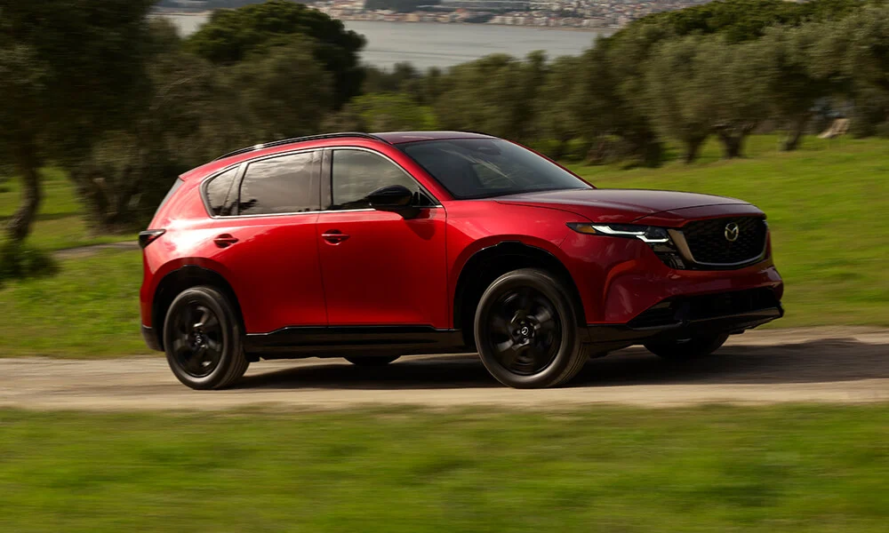 Vue latérale du Mazda CX-5 rouge vibrant cristal métallisé roulant et dépassant un champ herbeux avec des arbres, sur un arrière-plan flou causé par le mouvement.